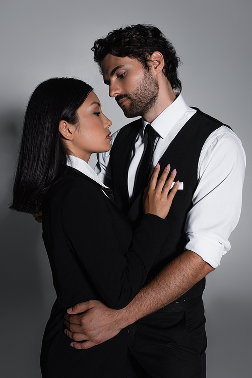 brunette man in formal wear hugging pretty asian woman on grey background