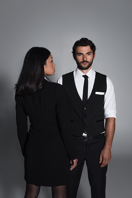 back view of asian woman in black blazer dress near stylish man looking at camera on grey background