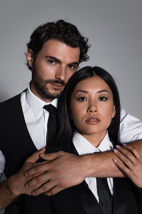 brunette man embracing sensual asian woman while looking at camera isolated on grey