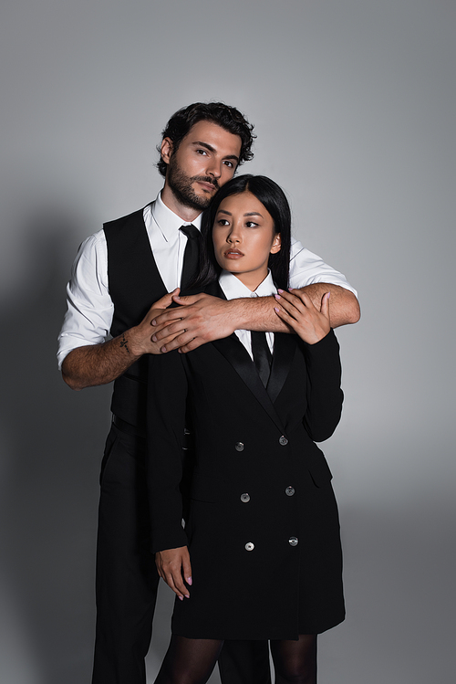 stylish man hugging asian woman in black blazer dress on grey background