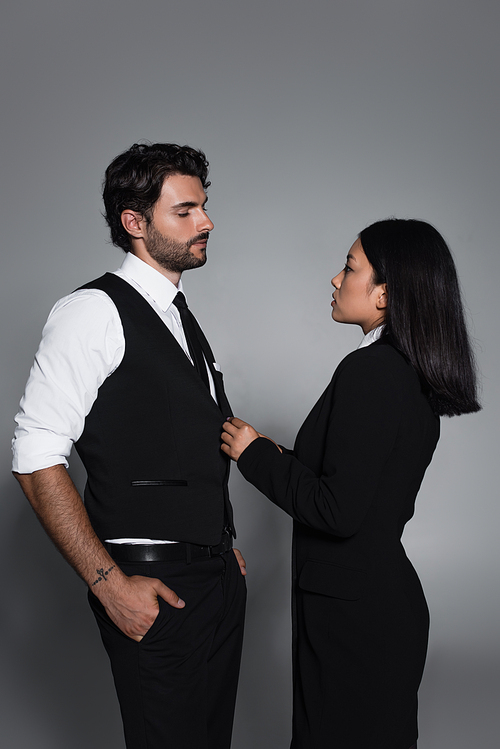 side view of elegant asian woman near stylish man standing with hands in pockets on grey background