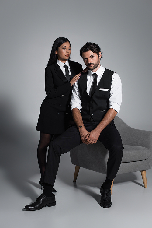 seductive asian woman in black blazer dress near stylish man sitting on armchair on grey background