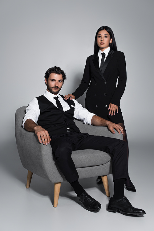 elegant man sitting in armchair near asian woman in black blazer dress on grey background