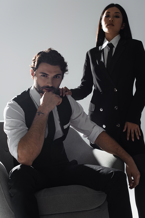 brunette stylish man sitting in armchair near elegant asian woman on grey background