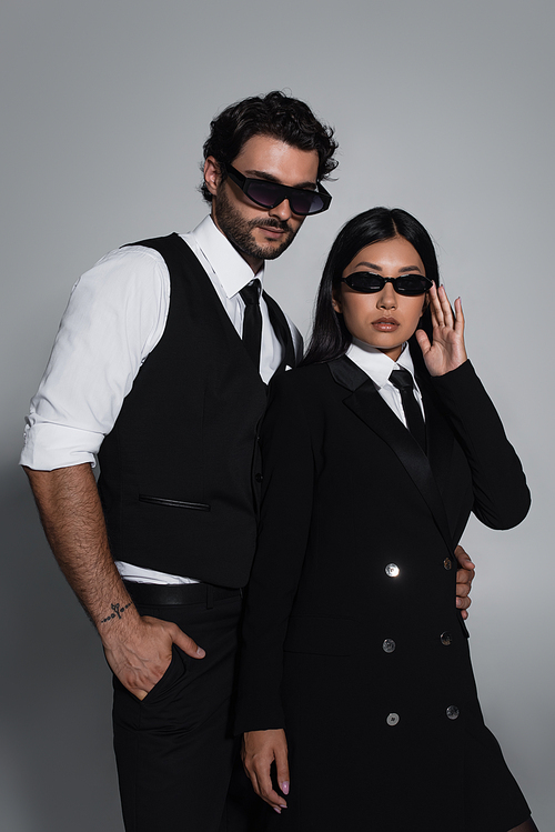 elegant interracial couple in formal wear and black sunglasses looking at camera on grey background