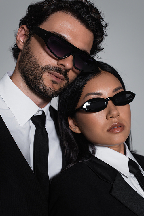 stylish interracial couple in formal wear and black sunglasses looking at camera isolated on grey