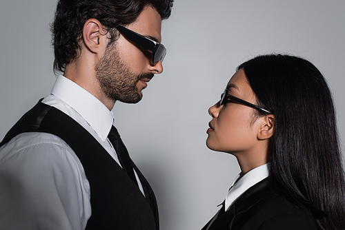 side view of interracial couple in black sunglasses looking at each other on grey background