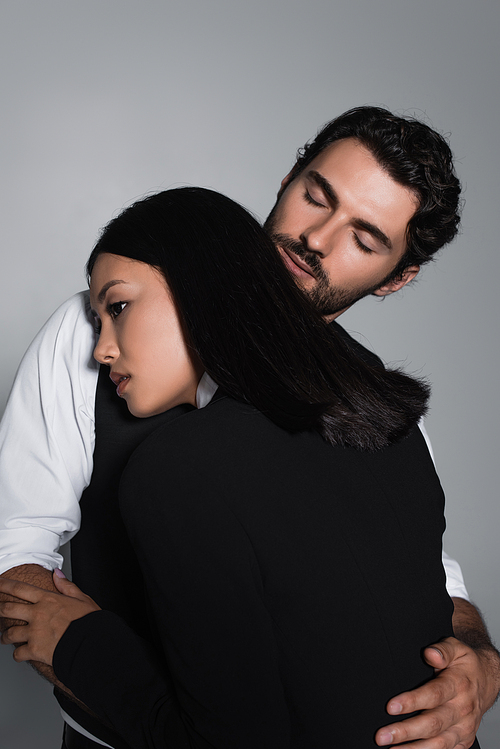 man with closed eyes hugging sensual asian woman isolated on grey