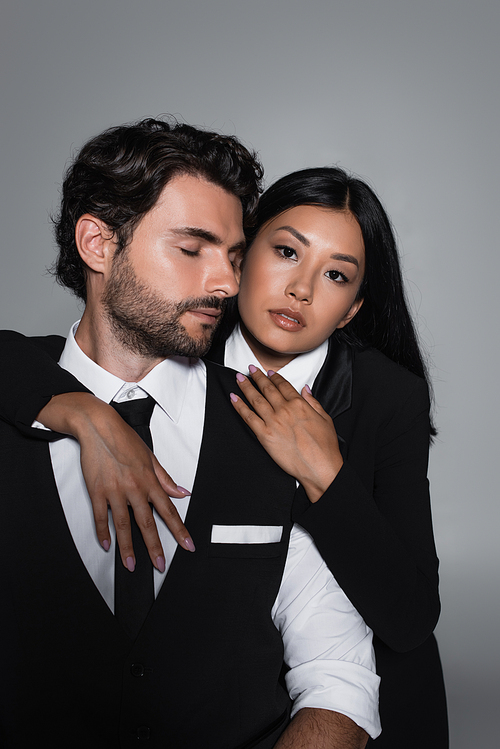 pretty asian woman embracing elegant man and looking at camera isolated on grey