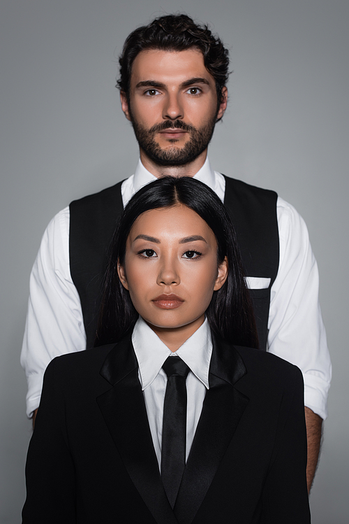 young asian woman and brunette man in formal wear looking at camera isolated on grey