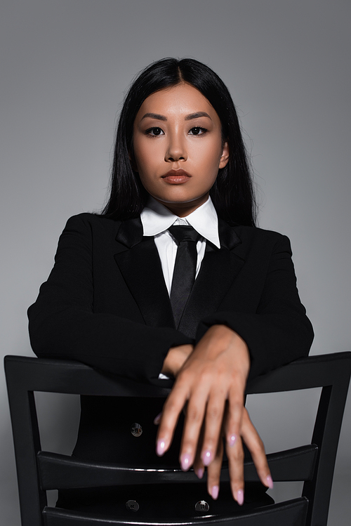 young asian woman in black formal wear looking at camera while posing on chair isolated on grey