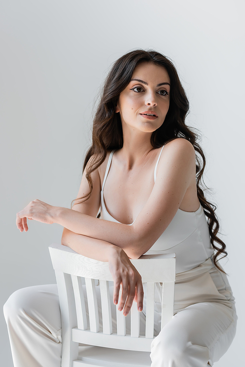 Brunette woman in white clothes sitting on chair isolated on grey