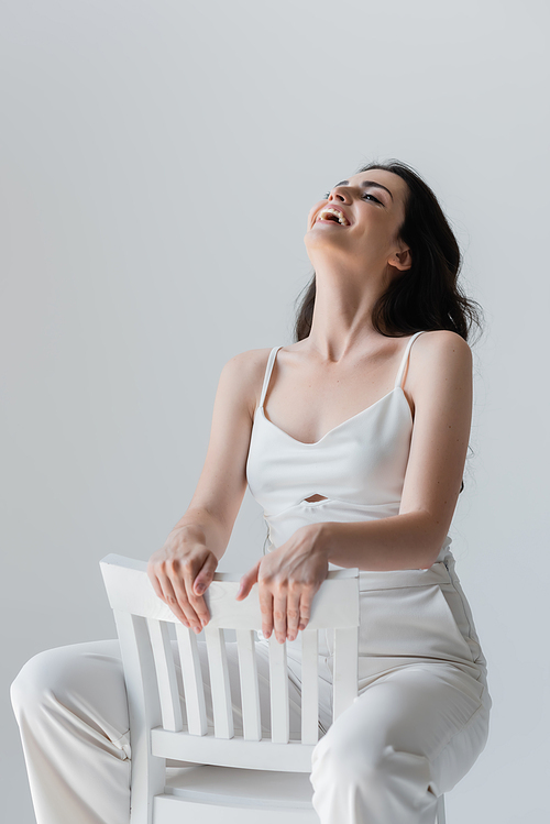 Positive brunette woman laughing while sitting on chair isolated on grey