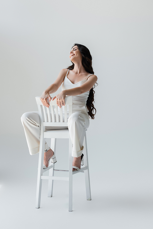 Full length of stylish brunette woman smiling while sitting on chair on grey background