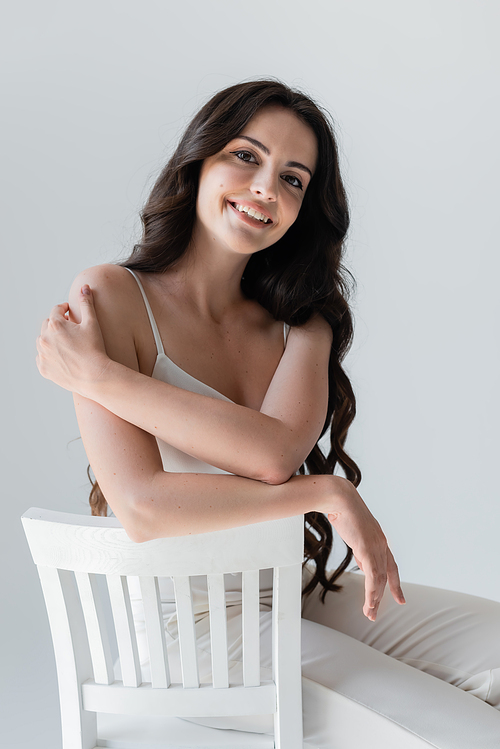 Positive long haired model in white clothes sitting on chair isolated on grey