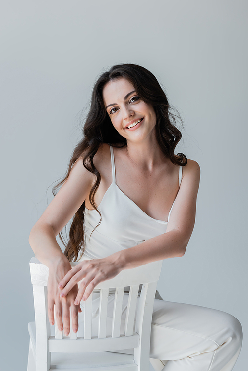 Happy brunette woman looking at camera while posing on chair isolated on grey