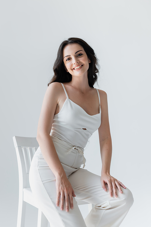 Pretty woman in white clothes smiling near chair isolated on grey
