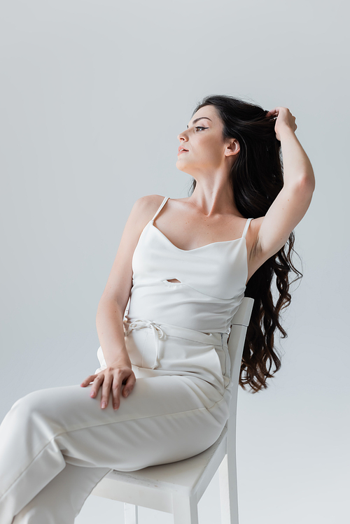 Young model in white clothes touching long hair while sitting on chair isolated on grey