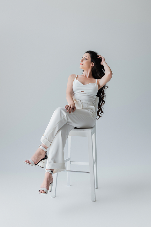 Full length of young model in white clothes and heels sitting on chair on grey background