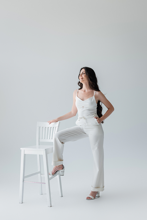 Smiling woman in white clothes posing near chair on grey background
