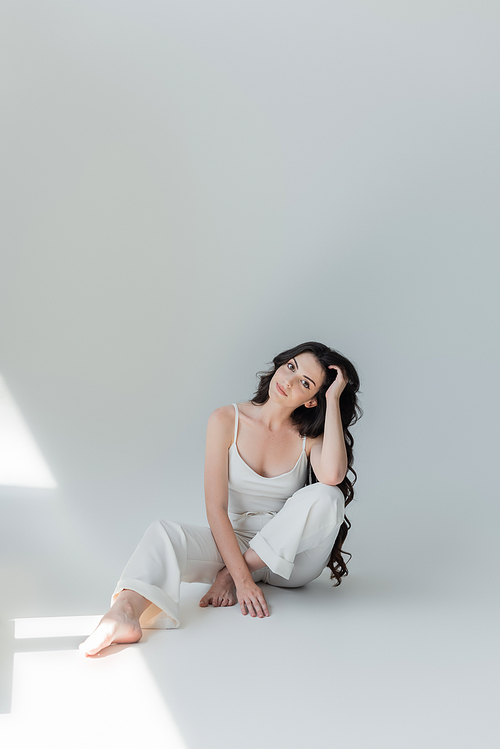 Long haired and barefoot woman in white clothes looking at camera on grey background