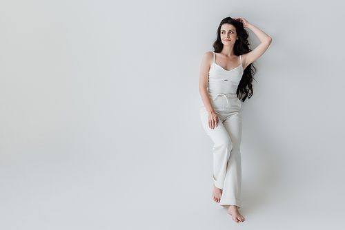 Full length of barefoot woman in white clothes smiling on grey background