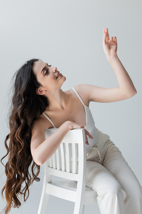 Stylish long haired woman gesturing while sitting on chair isolated on grey