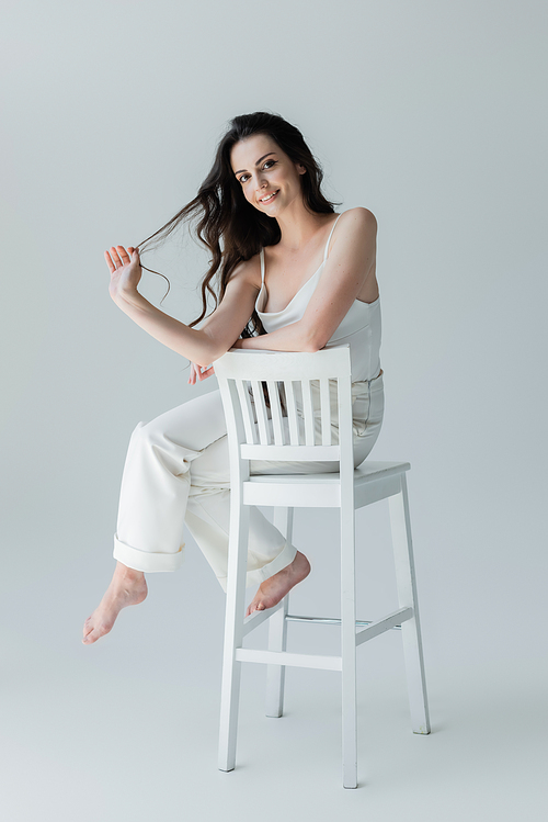 Full length of positive barefoot woman sitting on chair on grey background