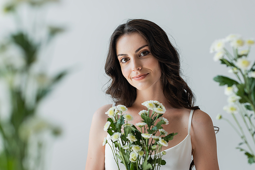 Smiling woman in white top looking at camera near blurred flowers isolated on grey