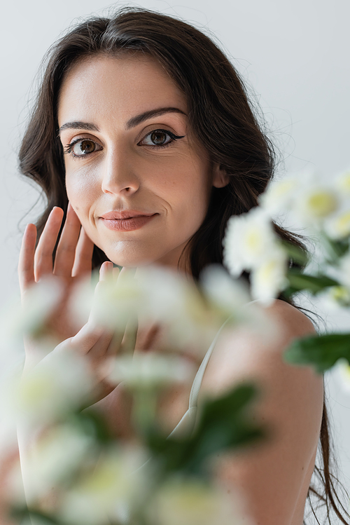 Portrait of brunette model with makeup looking at camera near blurred flowers isolated on grey