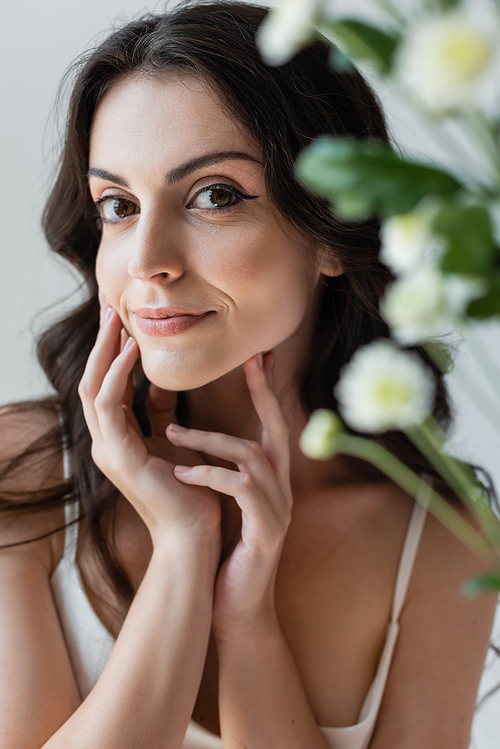 Brunette model with visage smiling at camera near blurred flowers isolated on grey