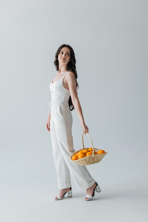 Full length of stylish model holding basket with oranges on grey background