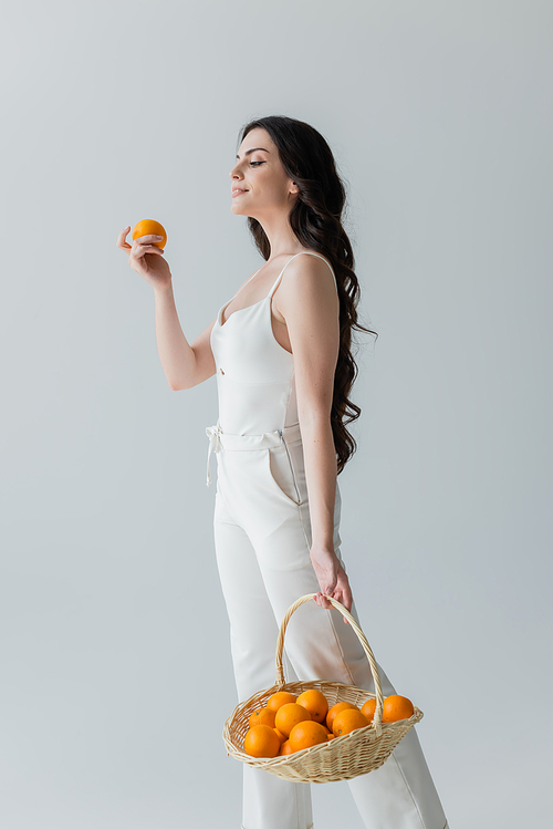 Smiling woman in white clothes holding basket with fresh oranges isolated on grey