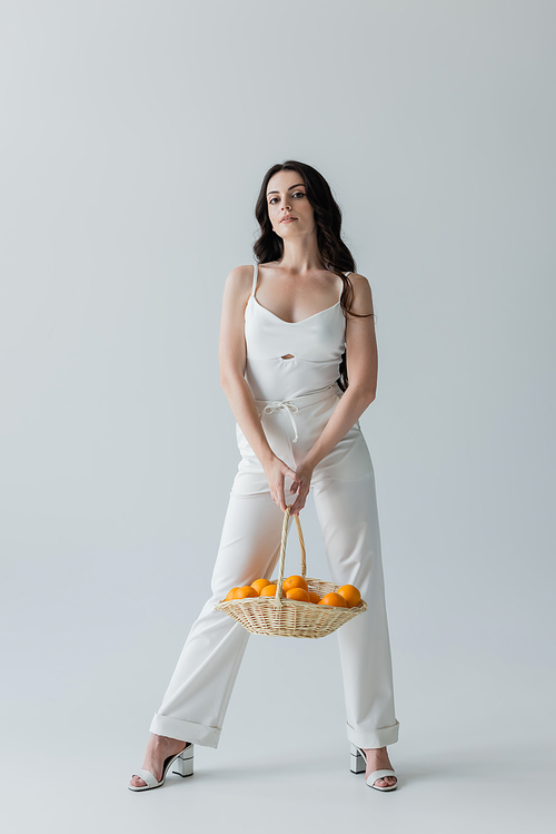 Full length of trendy woman holding basket with oranges on grey background