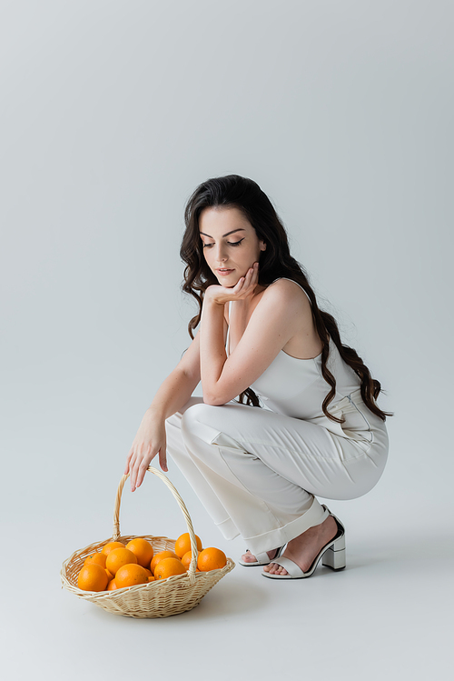 Long haired model in white clothes looking at basket with oranges on grey background