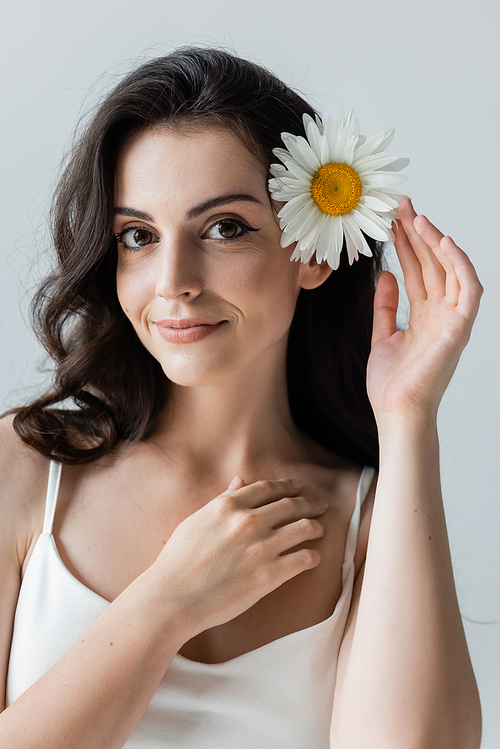 Portrait of smiling woman with makeup touching chamomile in hair and looking at camera isolated on grey