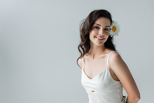 Happy brunette woman with chamomile in hair looking at camera isolated on grey