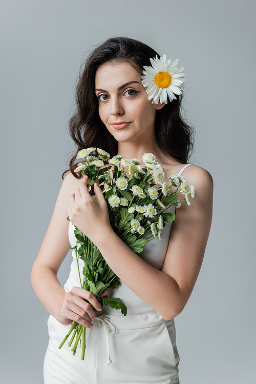 Pretty young woman holding bouquet of chamomiles while standing isolated on grey