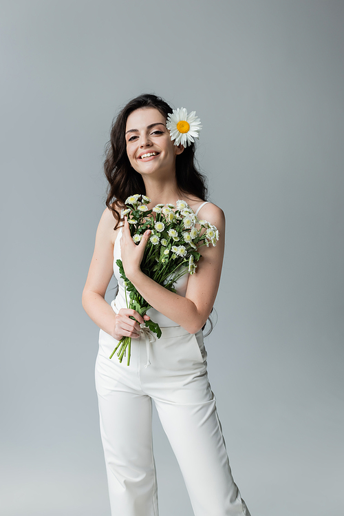 Positive woman in white clothes holding bouquet of chamomiles isolated on grey