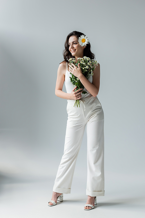 Full length of trendy woman in white clothes and heels holding chamomiles on grey background