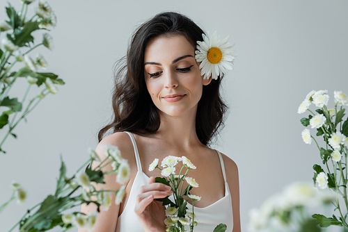 Smiling woman in white top touching flowers isolated on grey