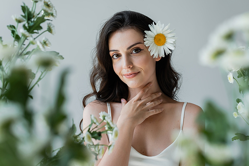 Portrait of smiling woman with chamomile in hair touching neck isolated on grey