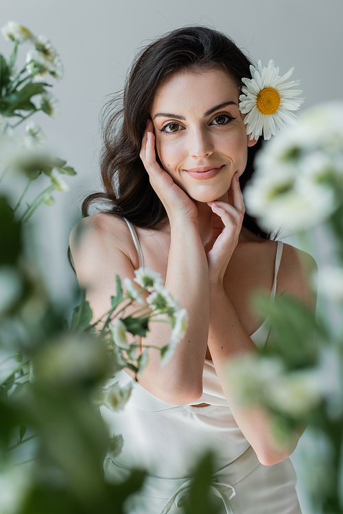 Pretty woman with chamomile in hair looking at camera near blurred flowers isolated on grey