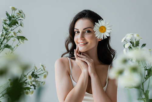 Pretty brunette woman with flower in hair smiling at camera near blurred flowers isolated on grey
