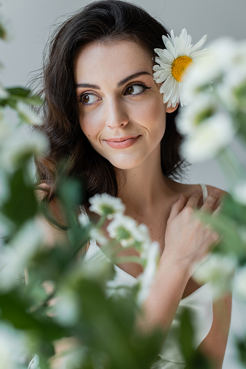 Smiling woman with visage and chamomile in hair looking away near blurred plants isolated on grey