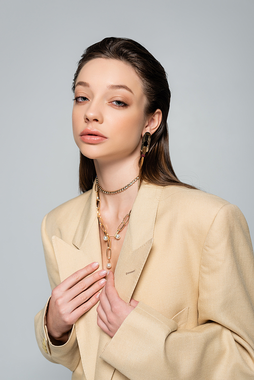young woman in beige blazer posing and looking at camera isolated on grey