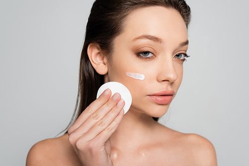 young woman with bare shoulders and cosmetic product on face holding cotton pad isolated on grey