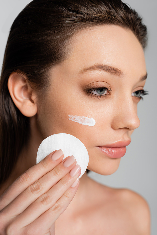close up of young woman with cosmetic product on face holding cotton pad isolated on grey