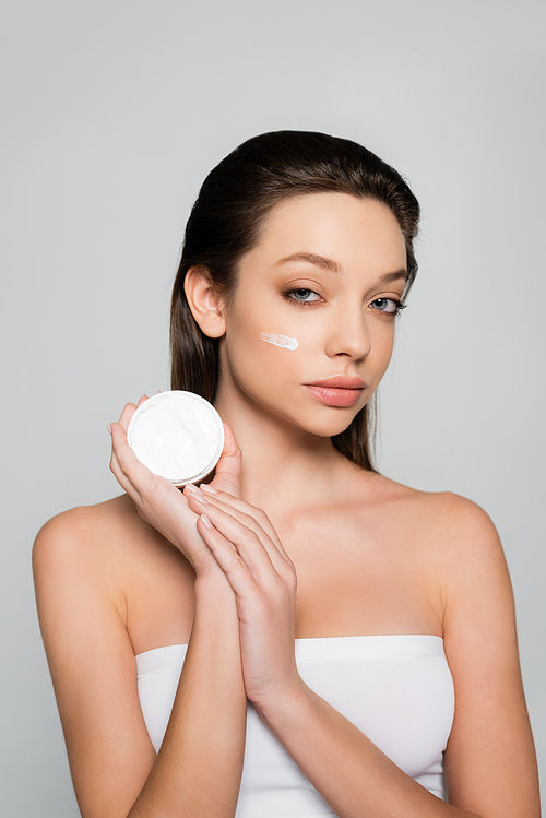 young woman with bare shoulders and cosmetic cream on cheek holding container isolated on grey