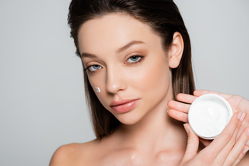 brunette woman with bare shoulders and cosmetic cream on cheeks holding container isolated on grey